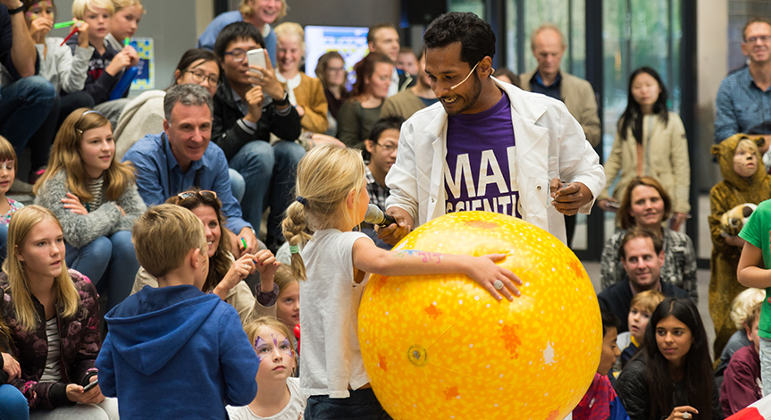 A girl in front of a large audience is holding a big yellow ball while speaking to a mad scientist holding a microphone